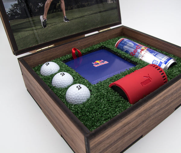Custom wooden box packaging with hinged lid displaying branded golf balls, a Red Bull energy drink, a Red Bull notepad, and a red Puma golf accessory. The interior is lined with synthetic green grass, providing a sporty and luxurious look.
